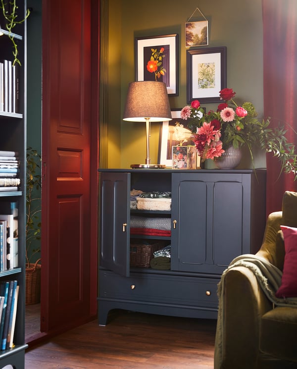 Cosy corner with SKOTTORP red lamp, dark cabinet, floral vase, art frames, and green chair.