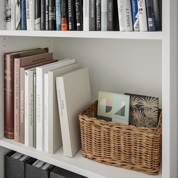 White bookshelf with woven basket holding colourful books, stacked books, and black-spined books beneath.