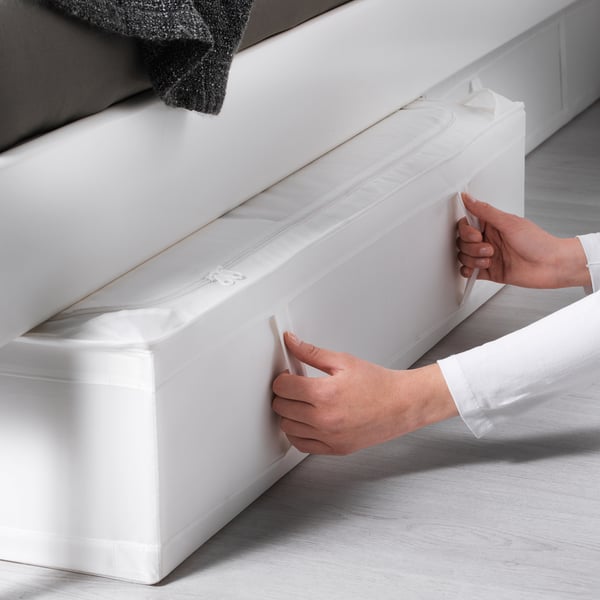 A person is pulling out a white SKUBB storage case from under a bed, showcasing its easy-access handles and dust-protective design.