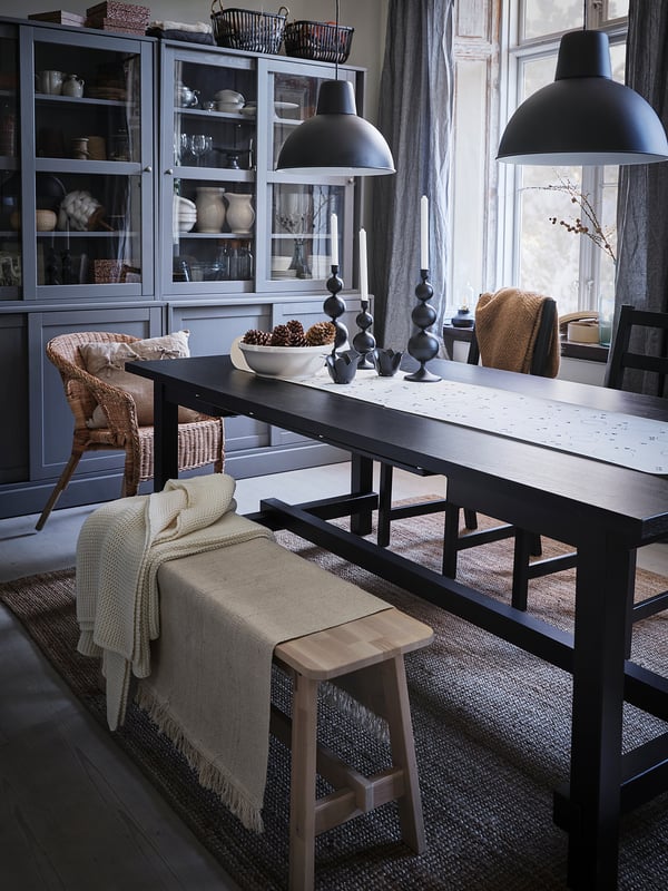 Modern dining area with black SKURUP pendant lamps, a long table, wooden chairs, blankets, and decorative items.