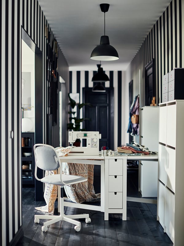 Modern home office with black and white decor, featuring a white desk, gray chair, sewing machine, and a SKURUP black pendant lamp.