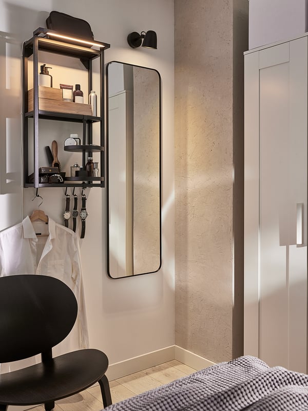 Modern bedroom featuring a large floor-length mirror, sleek black chair, led lit shelves with personal items, and a closet with a chequered blanket.