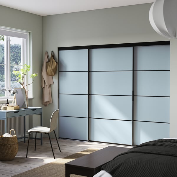 This image a modern, light grey sliding closet, SKYTTA, with black frames, mounted on a wall in a room with a desk and chair.