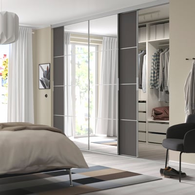 Modern bedroom with SKYTTA mirrored wardrobe, beige walls, wood floor. Tall wardrobe with clothes, black chair. Bed with beige cover.
