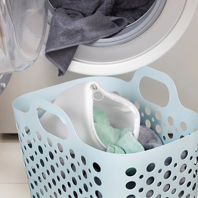 Blue laundry basket with grey clothes next to washing machine, mesh bag with green delicates.