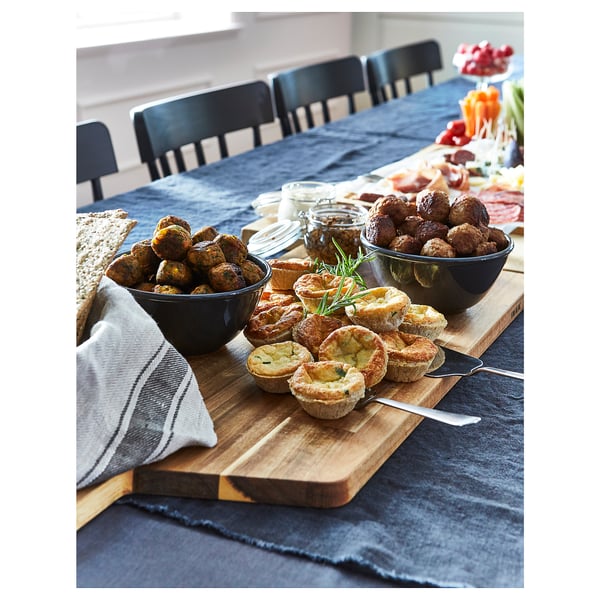 Black bowls with food on a wooden cutting board, placed on a grey table with black chairs around.
