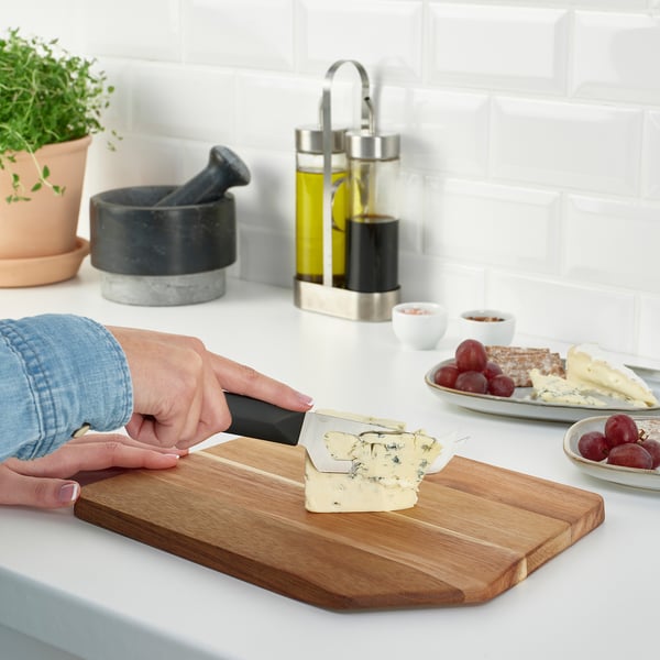 Person slicing cheese on a wooden cutting board on a kitchen counter. Nearby are grapes, crackers, and condiments.