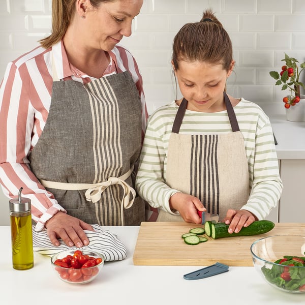Person and child prepare food, wearing aprons. Child uses kid-safe knife to cut cucumber.