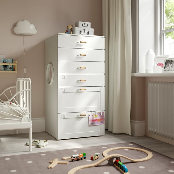 Kids room with white SMÅSTAD chest, 7 drawers, wooden handles. Near white bed, window with beige curtains, toys, and rug with polka dots.