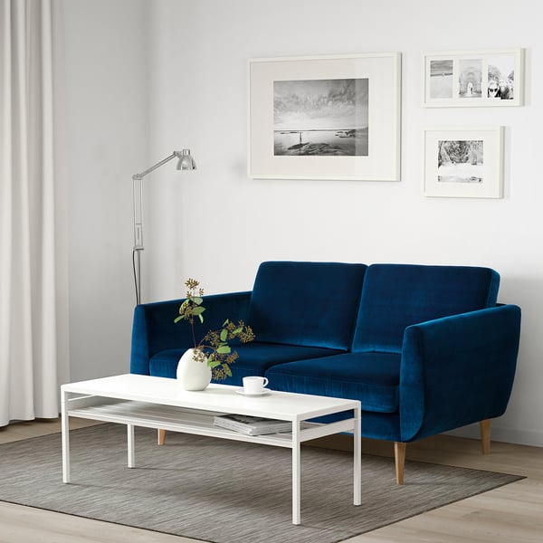 Modern living room with blue velvet SMEDSTORP sofa, white coffee table, and floor lamp. Artwork on white walls, plants, and curtains.