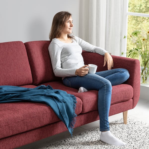A person sits on a red SMEDSTORP sofa, holding a cup, enjoying modern comfort with soft, resilient cushions and retro-style, cone-shaped legs.