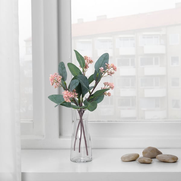 A clear vase with pink flowers and green leaves sits on a white windowsill, next to smooth pebbles. Bright sunlight streams in through the window.