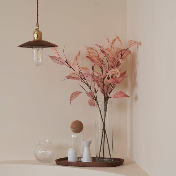 Rustic light hangs, illuminating pink leaves in tall vase, with candle holder and small vases on wooden tray.