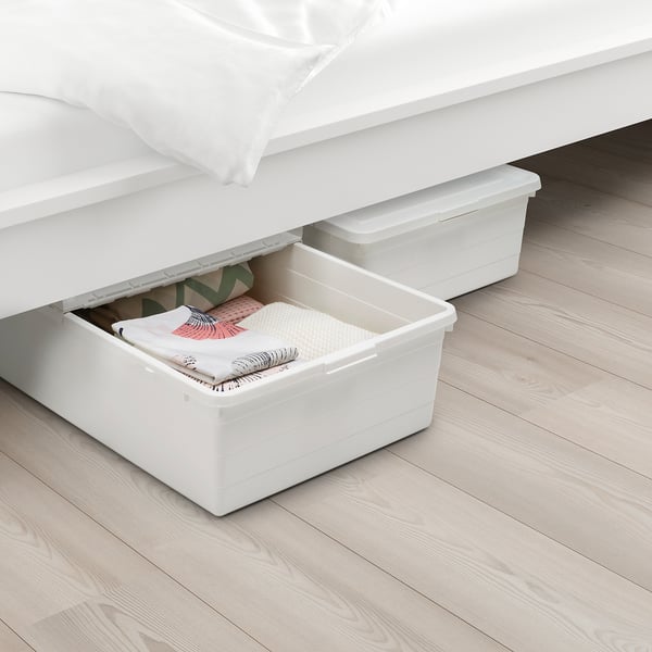 White plastic storage box under a bed, partially pulled out, holding folded clothes. Lid has small ridges for air circulation.
