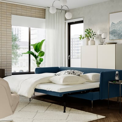 Modern bedroom with blue SÖDERHAMN sofa bed, white linens, plants, and large windows.