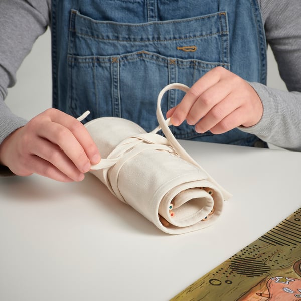 Person rolls up SOLFÅGEL pencil case with multiple pencils inside, demonstrating its portable and protective design.