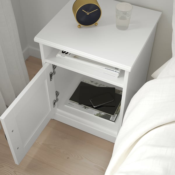 White bedside table with open drawer revealing laptop and charger. Top holds a gold clock and a water glass.
