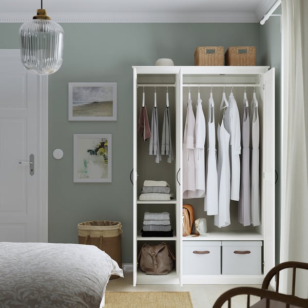 Neat bedroom: white wardrobe with mirror, clothes, shelves. Light blue wall, art, woven baskets, lamp.