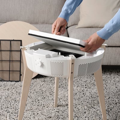 A person is placing a flat, white air philtre into a compact, white air purifier on wooden legs.