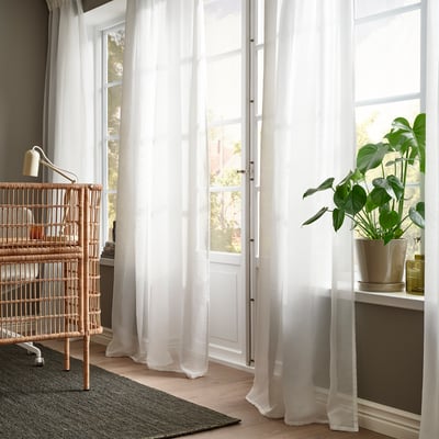 White curtains by tall window soften light, with wicker bookshelf holding books and plant alongside.