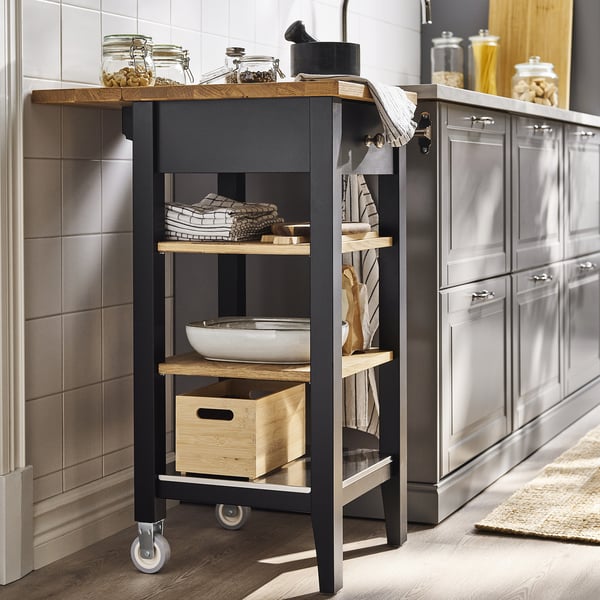 Black & wood kitchen trolley with 3 shelves & 4 wheels, stocked with jars, towels, & box.