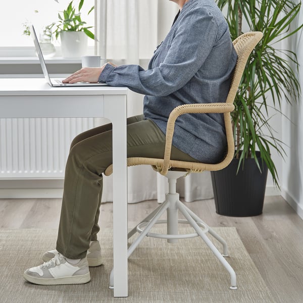 Person sitting on STIGBYGEL swivel chair, typing on laptop at white desk with green plant nearby.