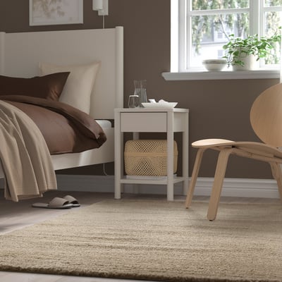 Bedroom with white bed, brown sheets, white rug, white bedside table, and wooden chair.