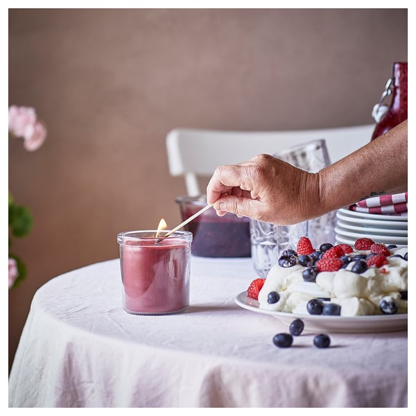 Person lights pink candle near berry dessert. Table set with white cloth, plates, flowers, and bottle. Warm, inviting atmosphere.