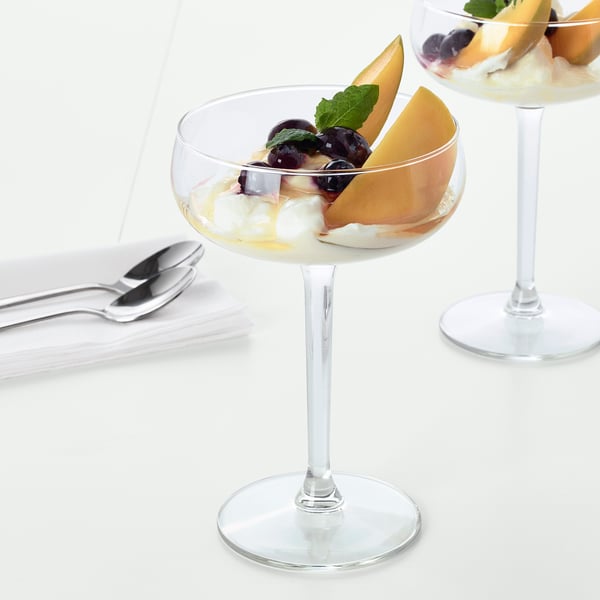 Two elegant, stemmed glasses filled with fruit and cream dessert, shown on a white table with spoons nearby.