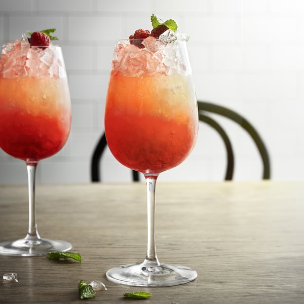 Two tall wine glasses filled with orange, reddish liquid, ice, raspberries, and mint leaves, placed on a wooden table.