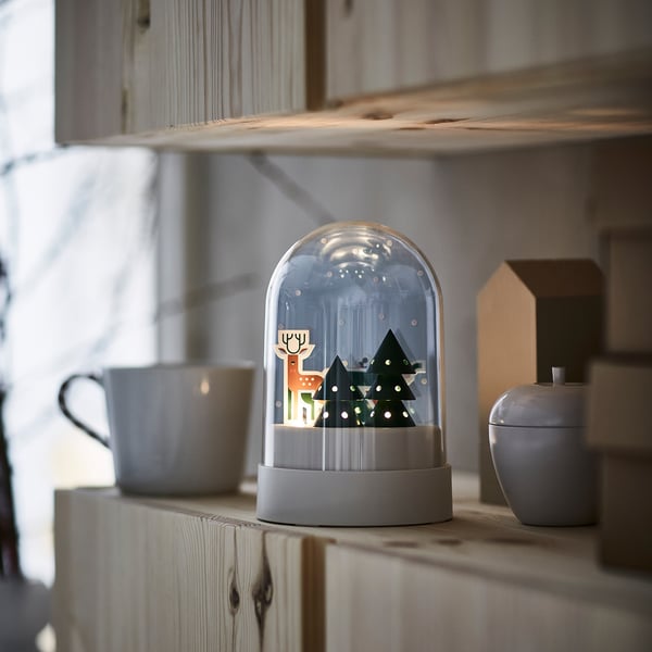 Snow globe lamp on shelf beside cups. Small, white, round, brightly lit inside. Trees & deer design.