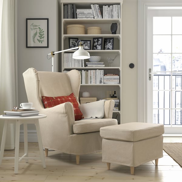 Cosy nook: beige armchair, ottoman, side table with cup & magazines. Large bookshelf behind, illuminated by a standing lamp. Door with window view.