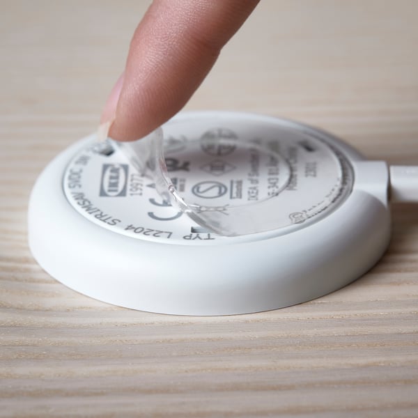 A hand pressing a round, white STRIMSÄV spotlight on a wooden surface. The spotlight illuminates when pressed, suitable for cabinets and closets.