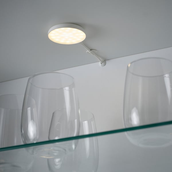 A modern, round spotlight mounted on a flexible arm illuminates a glass shelf with clear glasses, demonstrating its use for cabinet lighting.
