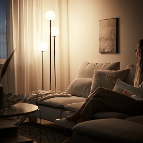 Cosy living room with grey couch, pillows, and lit floor lamp providing warm light. Person relaxes using a phone. Curtains draw soft light.