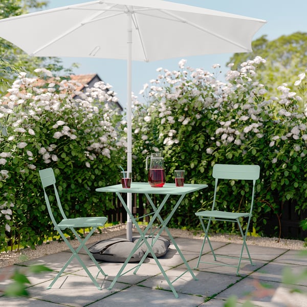 Serene patio scene with light green SUNDSÖ metal table and chairs, white umbrella, pink flowers, and drinks.