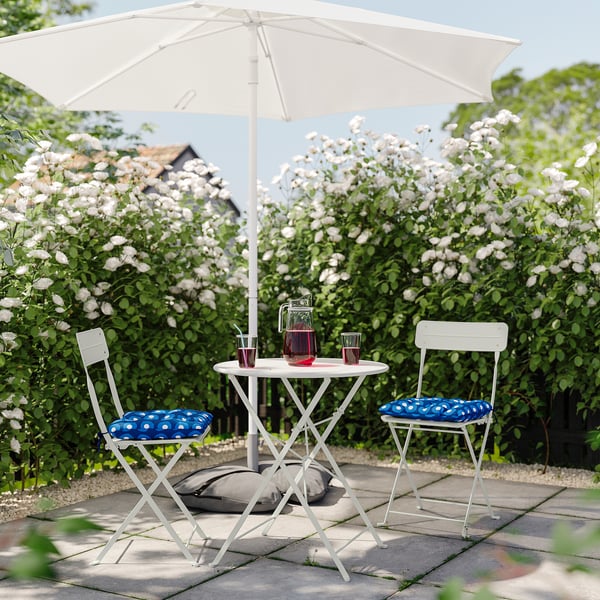 Small outdoor setup with white foldable table and chairs, blue cushions, white flowers, and a drink pitcher under a white umbrella.