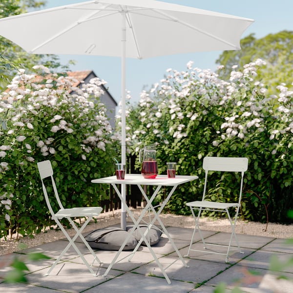 Small patio set with white folding table and chairs, red drinks, under white umbrella, surrounded by blooming bushes.