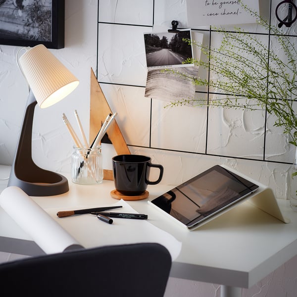 Modern workspace with white desk, black mug, tablet, lamp, pens, and plant.