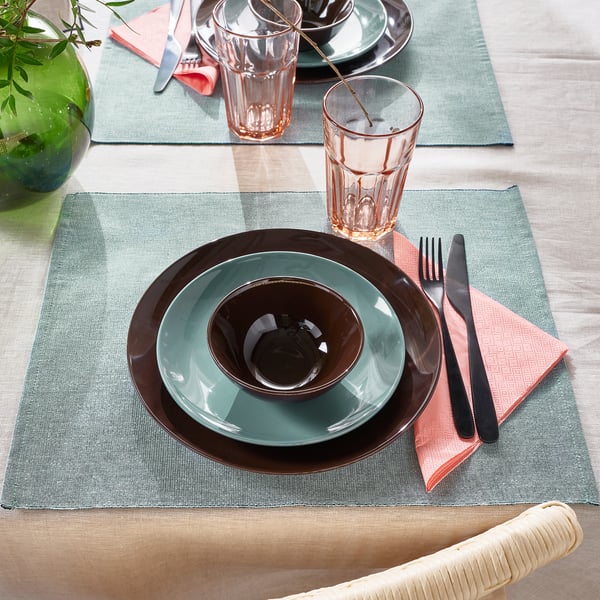 A table setting with brown and teal plates, pink glasses, and cutlery on green-blue placemats.