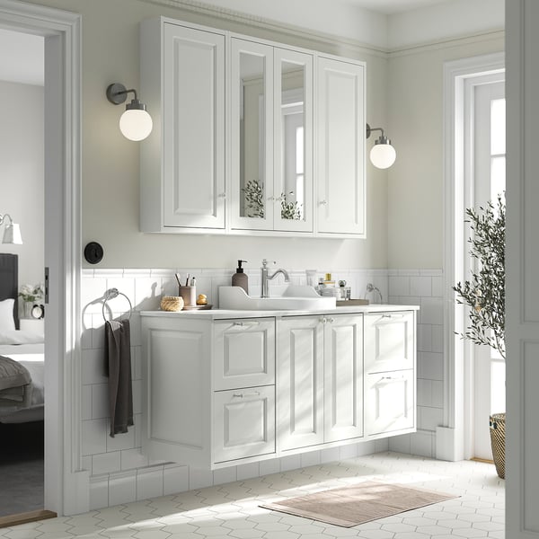 White bathroom vanity with two drawers, two doors, and a sink. Mirrors above, wall-mounted lights, grey towels, and a rug.