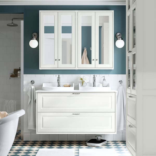 Modern white bathroom with large vanity unit, sink, and mirror cabinet. Floor has blue-gray tiles. Mounted towels, light fixtures, and accessories.