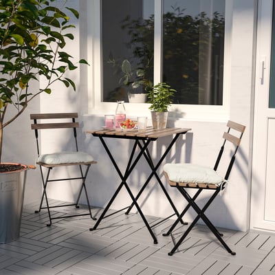 Small, black metal TÄRNÖ bistro set with acacia wood tops and cream cushions on a balcony, next to a white wall with a window and potted plants.