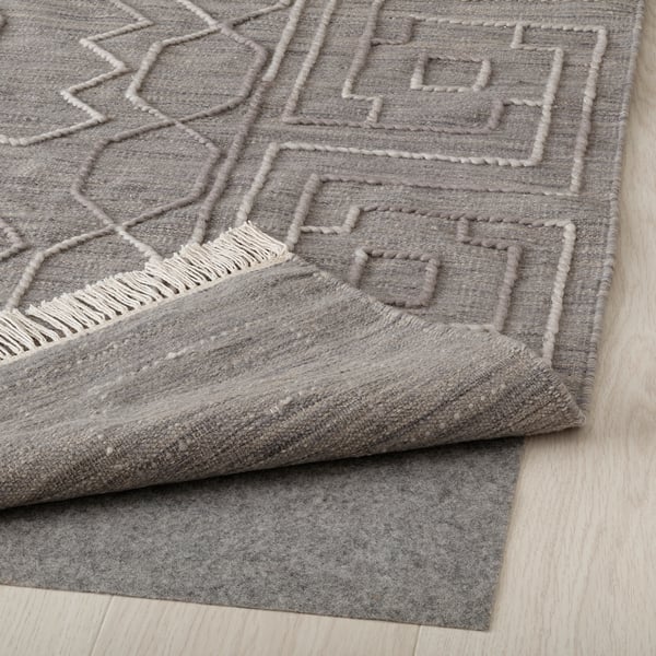 Grey, handwoven wool rug with geometric pattern lies on light wooden floor. Raised patterns and fringe detail visible.