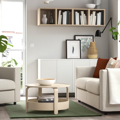 A cozy living room with a TIDTABELL light green rug, neutral furniture, cotton wool, coffee table, chairs, bookshelf, plants, and lamp.