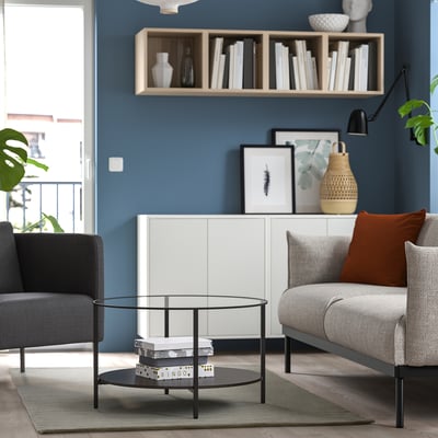 Round glass coffee table, black legs, central stack, grey rug, modern living room with grey sofa, orange pillow, blue wall, wooden floor.