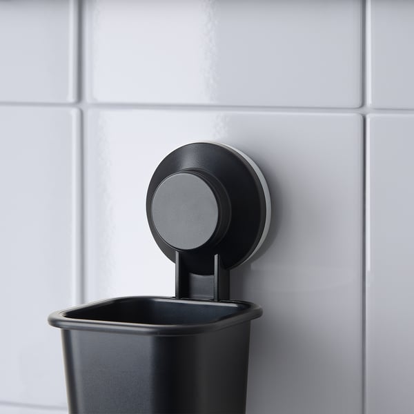 A black TISKEN toothbrush holder attached to a tiled wall. Its held by a strong suction cup, keeping the counter clean.