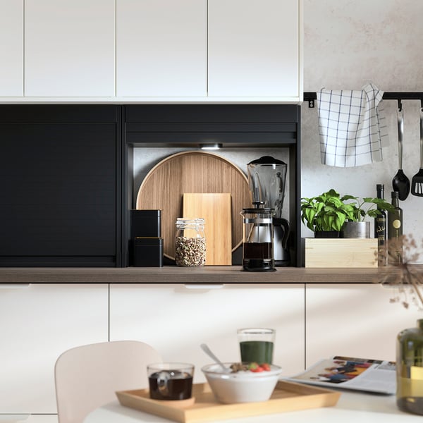 Modern kitchen with black TITTEBO cabinets, wooden countertop, and plants. White chairs and tray with drinks and fruit.