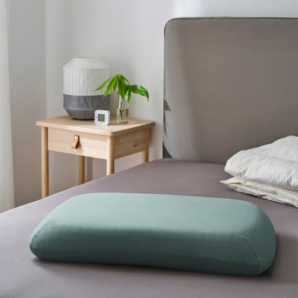 Bed with green pillowcase on purple sheet, next to wooden bedside table with lamp and plant.