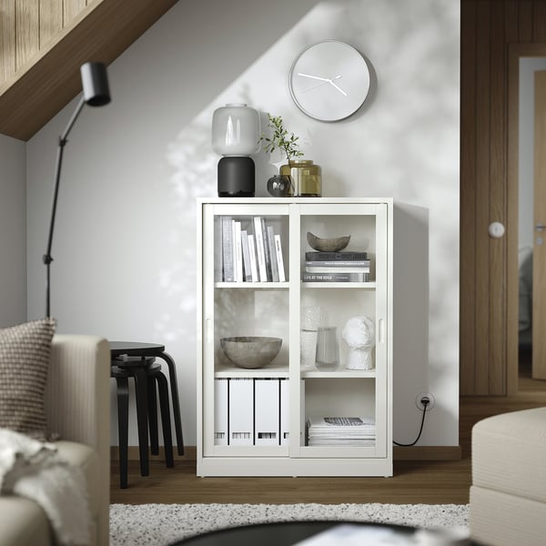 White wooden bookcase with sliding glass doors; holds books, vases, and a clock, in a modern living room.